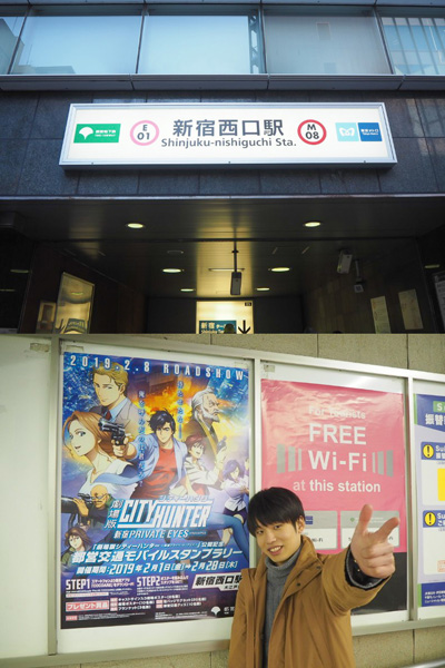 新宿西口駅に到着し、シティーハンターのポスター横で銃弾を撃つポーズを決める編集部員