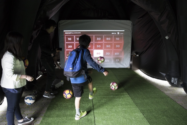 「Playbox」内でスクリーンに投影された点数表示に向かってサッカーボールを蹴る子ども